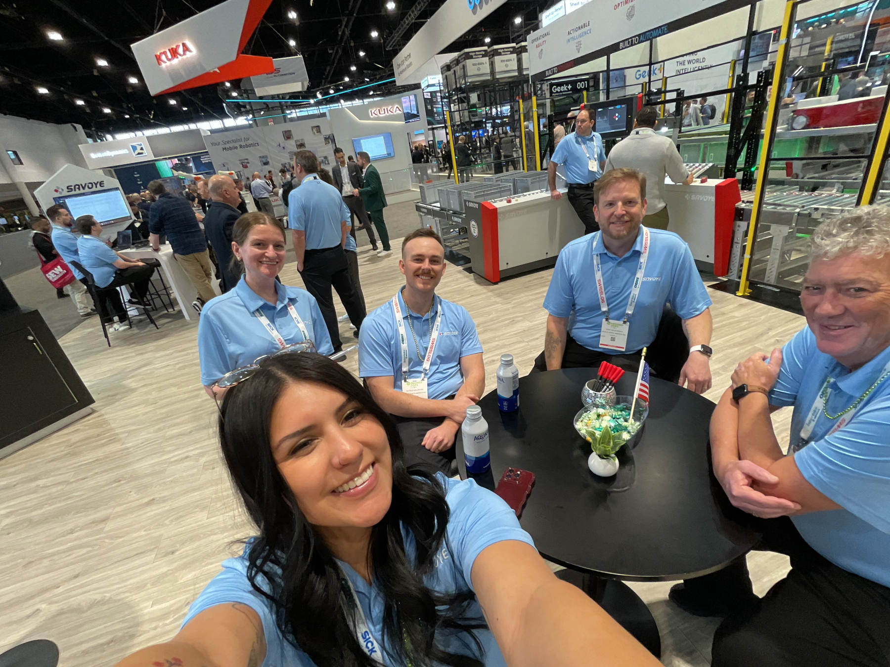 SAVOYE North America team members in branded shirts at an industry trade show booth surrounded by warehouse automation equipment and live system demonstrations